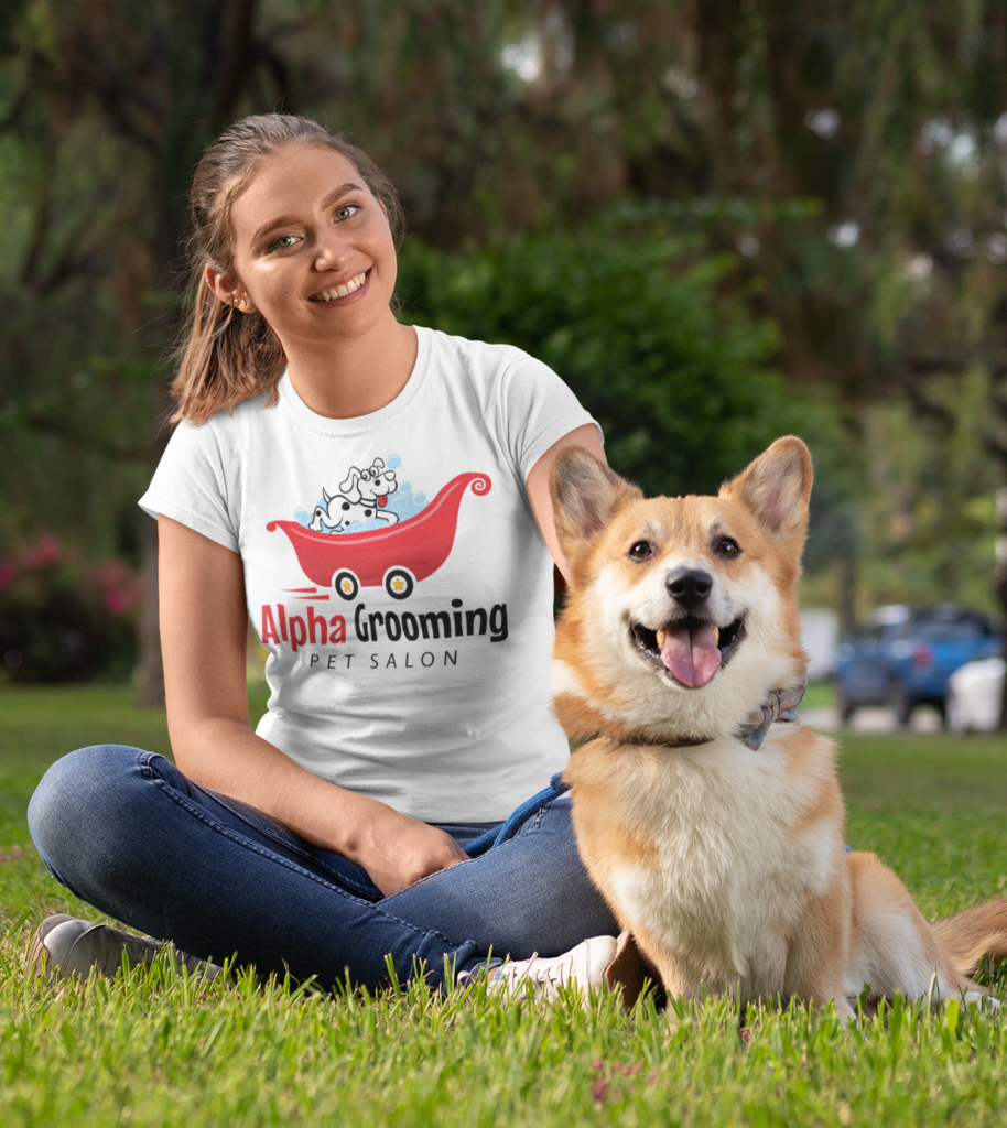 Woman sitting on grass with a smiling dog, wearing a white "Alpha Grooming Pet Salon" t-shirt; park setting with trees in the background—perfectly capturing the joy of mobile pet grooming.